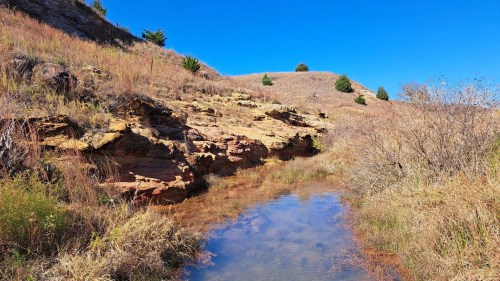 A serene landscape featuring a shallow stream surrounded by rocky terrain and grassy hills under a clear blue sky.