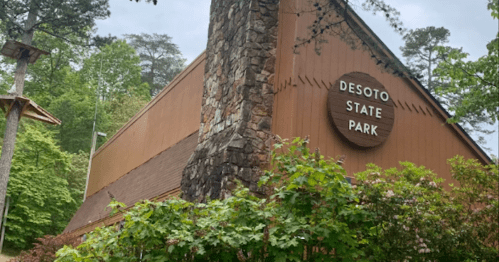 A wooden building with a stone chimney, featuring a sign that reads "Desoto State Park," surrounded by greenery.
