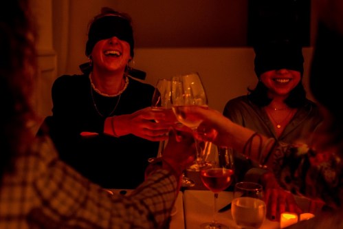 Two women wearing blindfolds laugh and toast with glasses of wine in a dimly lit setting.
