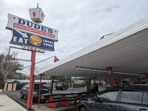 A retro diner sign for Dude's, advertising fresh food since 1965, with cars parked under a covered area.