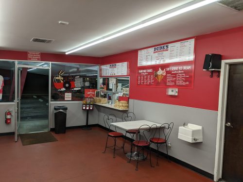 Interior of a casual eatery with red walls, a menu board, tables, and a counter displaying snacks and ice cream.