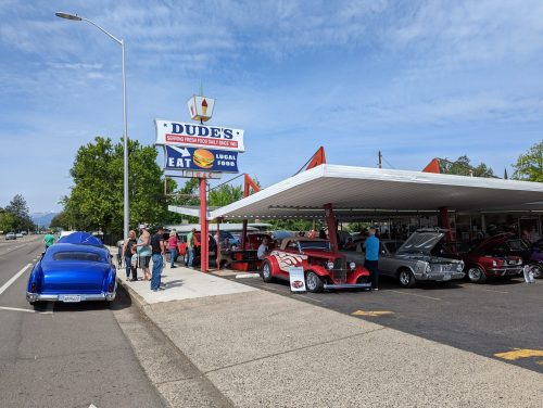 A classic car show outside Dude's diner, with vintage cars and a crowd enjoying the event under a clear blue sky.