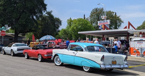 A row of classic cars parked outside a diner, with people gathered around on a sunny day.