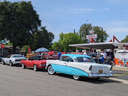 A line of classic cars parked outside a retro diner, with people gathered and an umbrella providing shade.