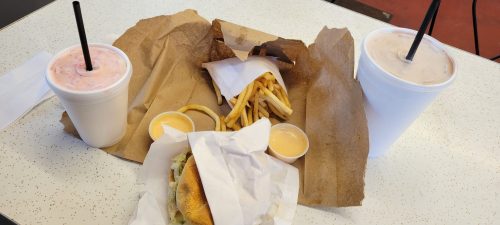 A table with two drinks, a burger wrapped in paper, fries, and dipping sauces on brown paper.