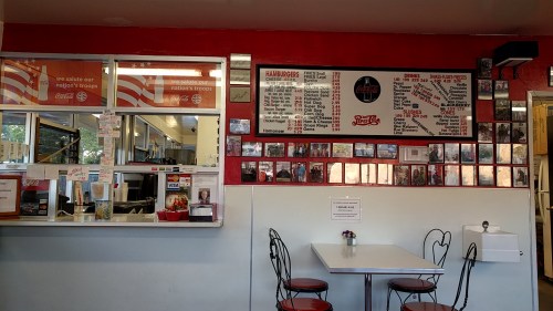 Interior of a diner with red walls, a menu board, and a small table with chairs. Photos decorate the wall.