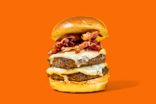 A stacked burger with two beef patties, melted cheese, and crispy bacon, set against a bright orange background.