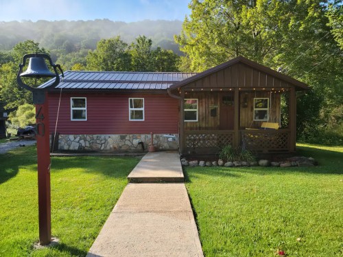 A charming red cabin with a stone foundation, surrounded by greenery and a clear blue sky. A pathway leads to the entrance.
