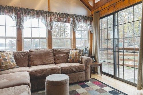 Cozy living room with a brown sectional sofa, colorful pillows, and large windows overlooking a deck and nature.