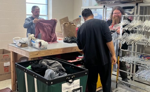 Two people are folding clothes at a table, while a third person sorts items from a bin in a clothing donation center.