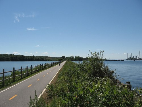 A scenic bike path alongside a calm river, with greenery and distant industrial structures under a clear blue sky.