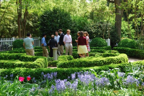 A group of people stands in a lush garden with neatly trimmed hedges and colorful flowers, engaged in conversation.