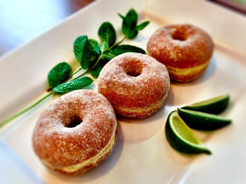 Three sugar-coated donuts on a white plate, garnished with mint leaves and lime wedges.