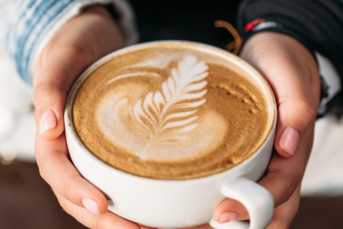 A person holds a white cup of latte art featuring a leaf design on the creamy surface.