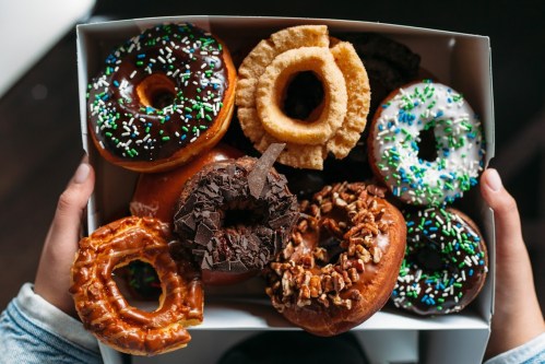 A box of assorted donuts, featuring chocolate, glazed, and sprinkled varieties, held by two hands.