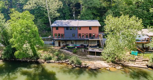 A two-story house by a river, surrounded by trees, with a deck and kayaks on the shore.