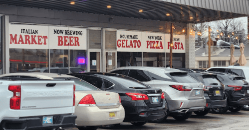 Exterior of an Italian market featuring signs for beer, gelato, pizza, and pasta, with parked cars in front.