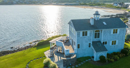 Aerial view of a coastal home with a patio, overlooking a shimmering bay and sandy beach in the background.