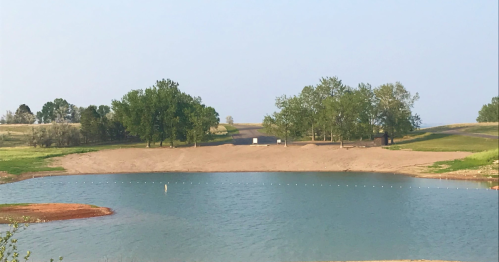 A serene landscape featuring a calm blue lake surrounded by sandy shores and green trees under a clear sky.