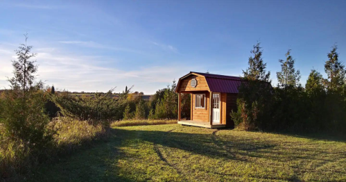 A small wooden cabin with a red roof sits on a grassy area, surrounded by trees and open fields under a clear blue sky.