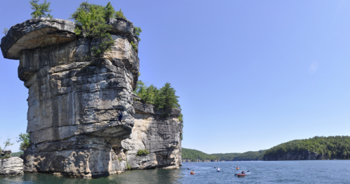 A large rock formation rises above a calm lake, surrounded by lush greenery under a clear blue sky.