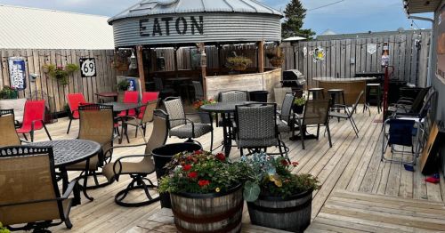 Outdoor seating area with tables and chairs, surrounded by wooden fencing and planters, featuring a rustic bar structure.