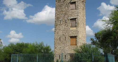 A tall stone tower with boarded windows, surrounded by greenery under a partly cloudy sky.