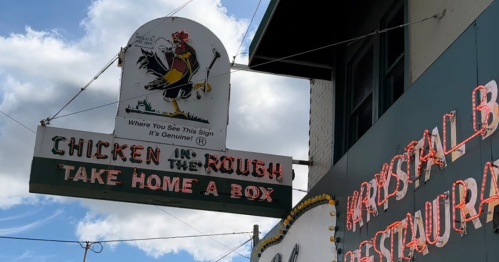 Sign for "Chicken in the Rough" restaurant featuring a rooster and the text "Take Home a Box" against a cloudy sky.