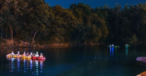 A group of people in kayaks paddles on a calm river at night, surrounded by trees and soft glowing lights.