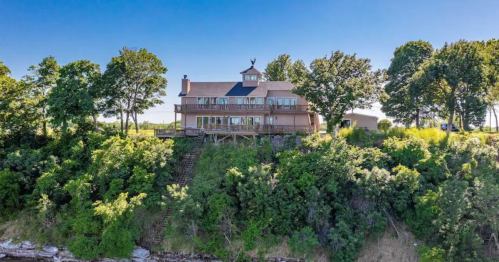 A large, two-story house on a hillside surrounded by trees, overlooking a body of water under a clear blue sky.