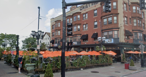 A lively outdoor restaurant with orange umbrellas, surrounded by greenery and a historic building in the background.