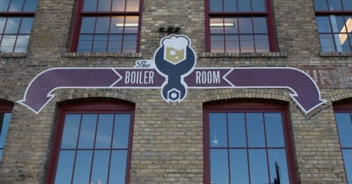 Sign for "The Boiler Room" with arrows pointing left and right, set against a brick building with large windows.
