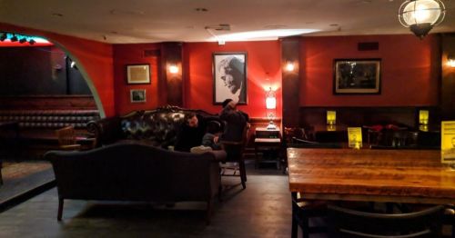 Cozy bar interior with warm lighting, a large wooden table, and a vintage portrait on the wall.