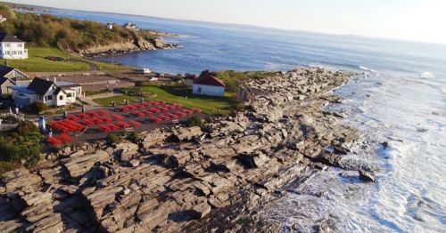 Aerial view of rocky coastline with a parking area and houses, overlooking the ocean on a sunny day.