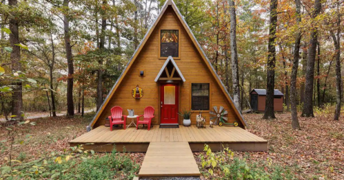 A cozy A-frame cabin surrounded by trees, featuring a red door, wooden deck, and two red chairs on the porch.