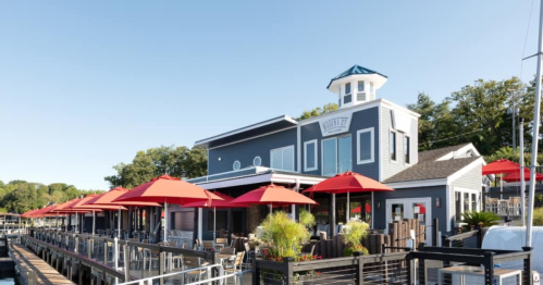 A waterfront restaurant with red umbrellas, a blue-roofed tower, and outdoor seating along a dock.