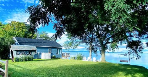 A serene lakeside view featuring a blue house, green lawn, and white chairs by the water under a clear blue sky.