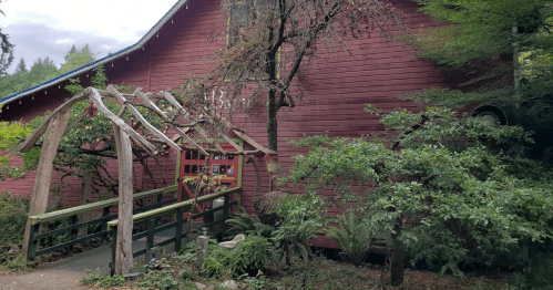 A red wooden building surrounded by greenery, featuring a wooden archway leading to a path.
