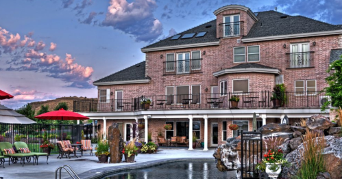A luxurious home with a pool, surrounded by greenery and patio furniture, under a colorful sky at sunset.