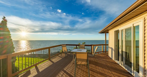 A sunlit deck overlooking a calm lake, with a table and chairs, surrounded by greenery and a clear blue sky.