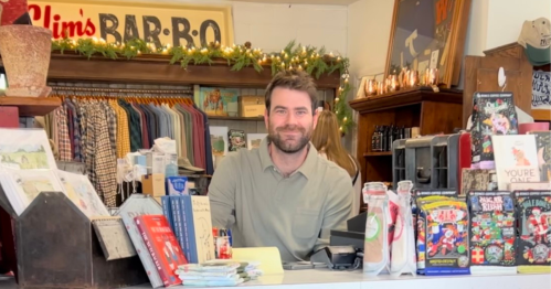 A smiling man stands behind a shop counter filled with books and merchandise, decorated for the holidays.