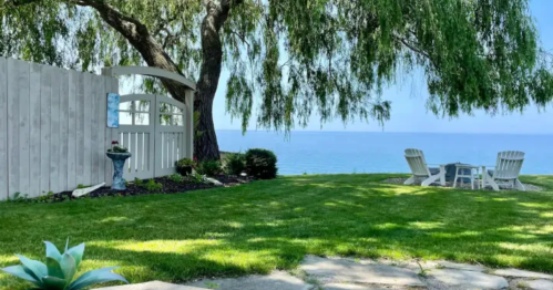 A serene lakeside view with a grassy yard, white chairs, and a gate under a willow tree.