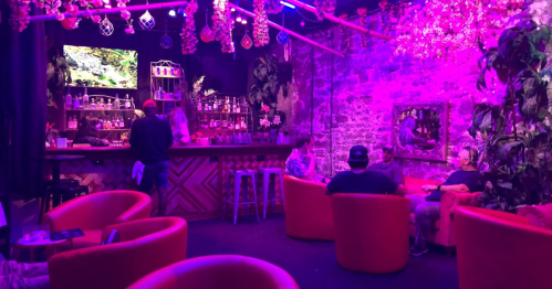 A vibrant bar scene with purple lighting, featuring colorful seating and patrons enjoying drinks in a lively atmosphere.