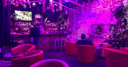 A vibrant bar scene with purple lighting, featuring colorful seating and patrons enjoying drinks in a lively atmosphere.