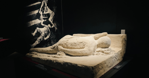 A sculpted figure lying down, displayed in a dark exhibit with a faint background image.