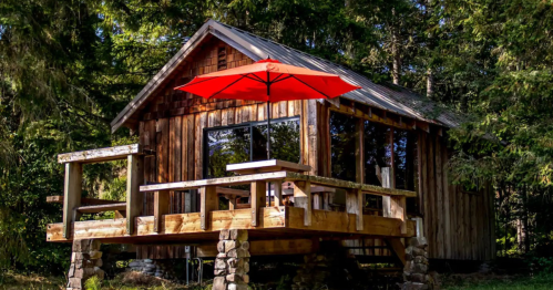 A rustic wooden cabin with a red umbrella on a deck, surrounded by lush greenery.