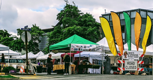 A farmers market setup with colorful flags, tents, and a "Road Closed" sign on a street corner.