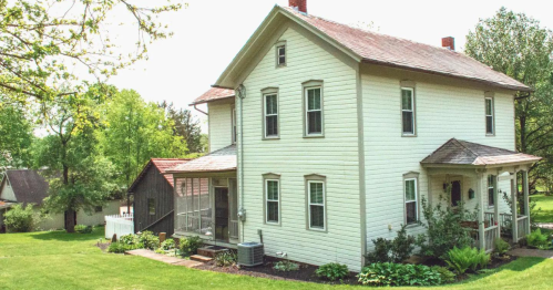 A two-story white house with a porch, surrounded by green trees and a well-maintained lawn on a sunny day.