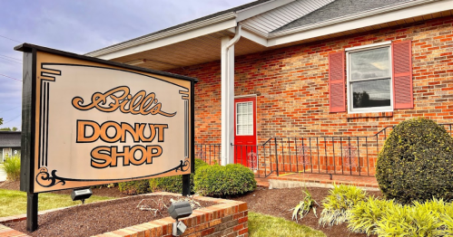 Sign for "Bill's Donut Shop" in front of a brick building with red shutters and a welcoming entrance.