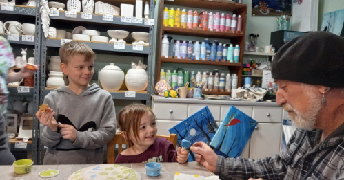 Two children and an older man are painting pottery in a colorful studio filled with art supplies.